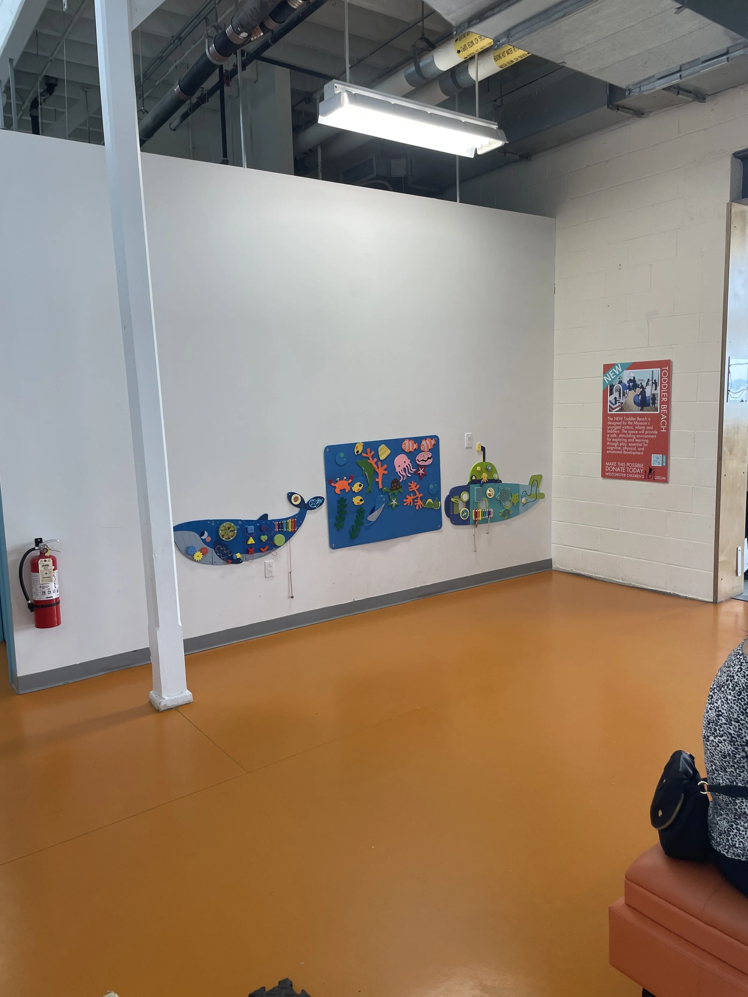 Interior view of a room with orange floor, white walls, and ceiling with exposed ducts and fluorescent light. On one wall, there are colorful decorations of a whale, a sea creature scene, and a submarine. A fire extinguisher is mounted on the left side. A person with a patterned shirt is sitting on an orange seat on the right, with a black bag beside them. A red poster is displayed on the wall to the right.