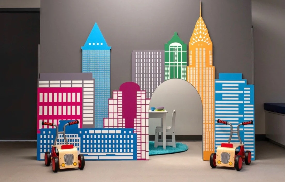 Colorful cardboard cutouts of city skyscrapers with two red ride-on toy cars and a small table and chair in the center, creating a children's play area.