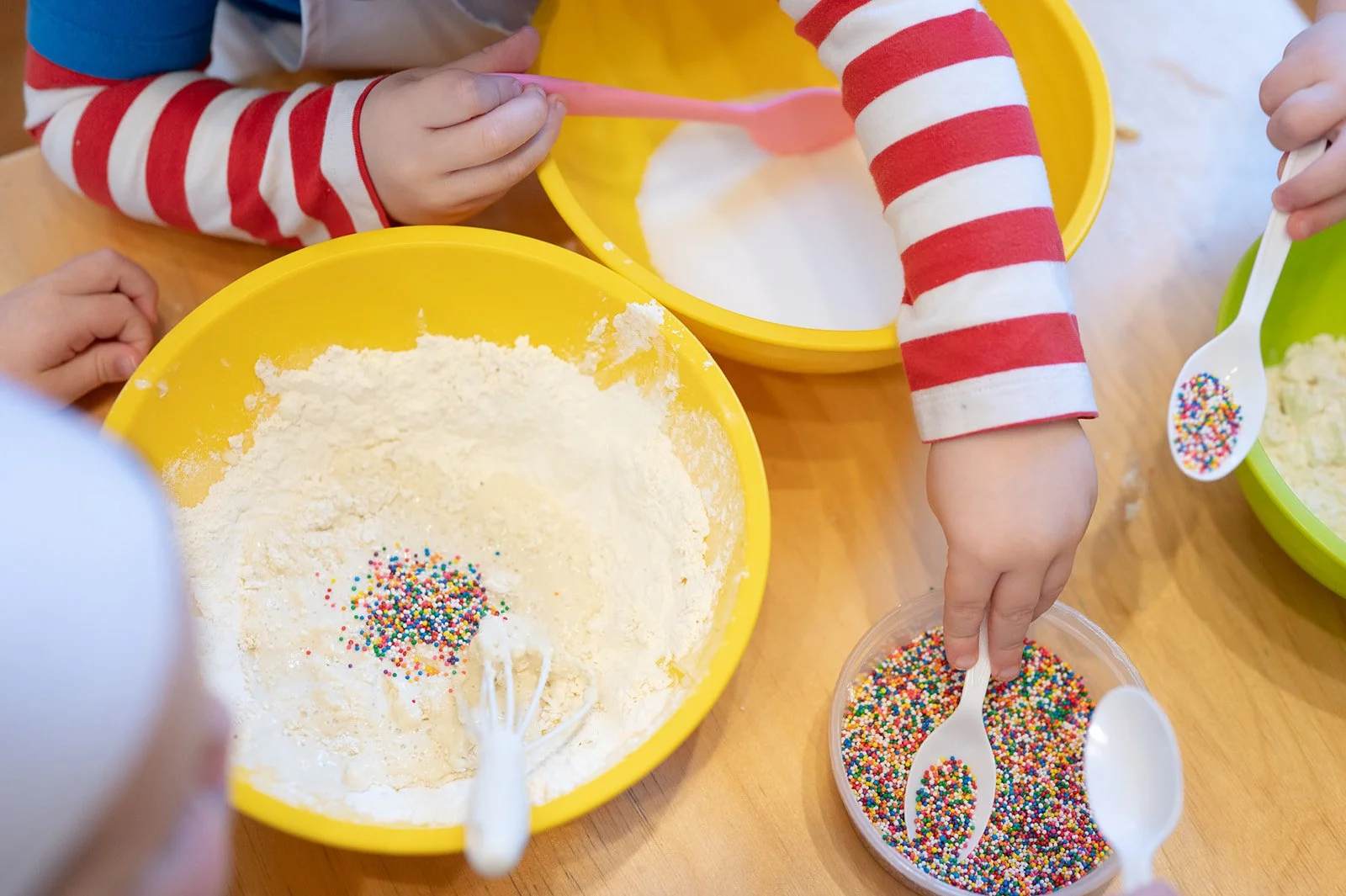 Children's hands mixing cake ingredients with rainbow sprinkles in bowls, wearing red and white striped sleeves.