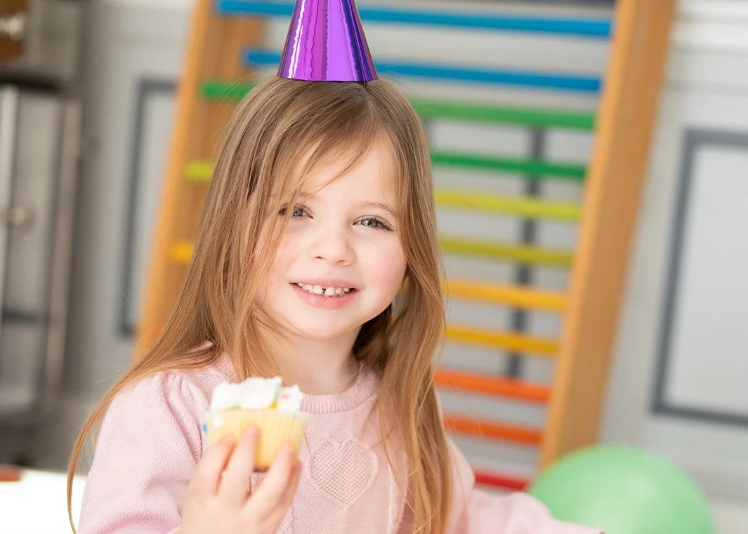 A young girl with long red hair and a pink shirt, wearing a purple party hat, smiling and holding a cupcake with white frosting. In the background, there is a colorful wooden ladder and a green balloon.
