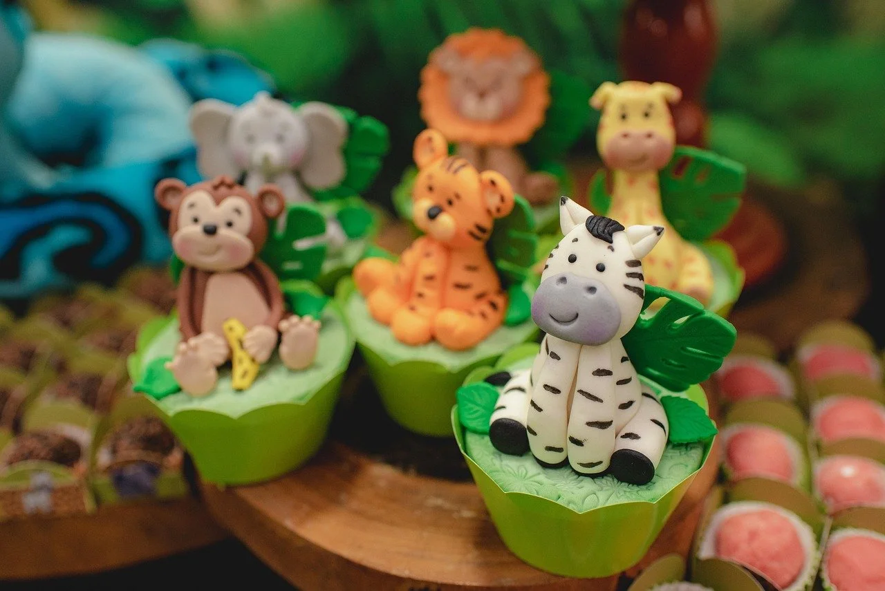 Cute animal-themed cupcakes with fondant figurines of a zebra, tiger, monkey, lion, and giraffe on a wooden surface.