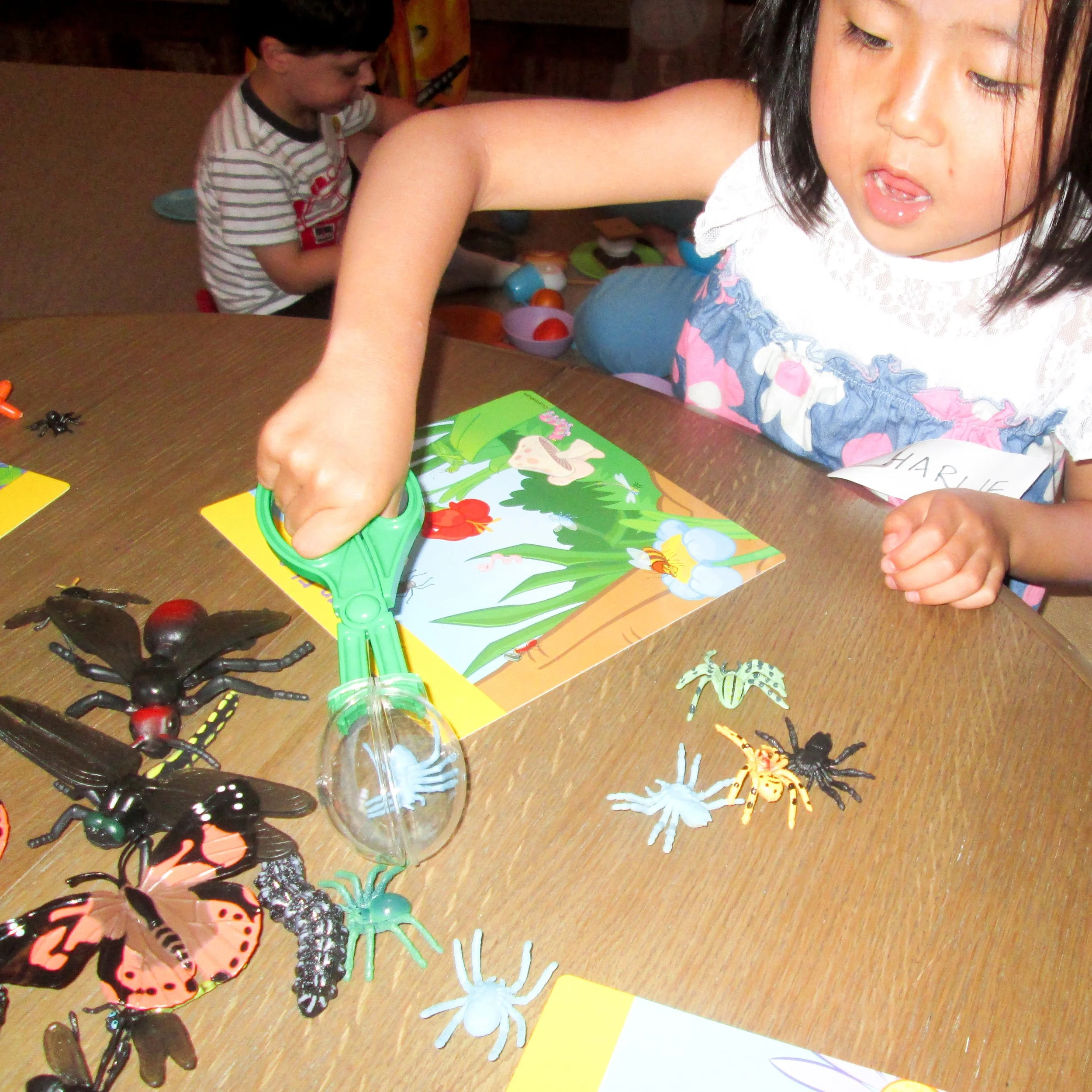 A young girl with a name tag labeled "HARMIE" playing with toy insects and a magnifying glass at a wooden table, with a boy in a striped shirt in the background.