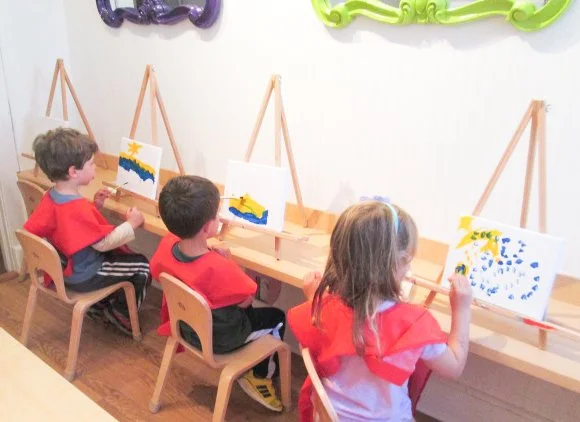 Three children sitting on chairs at a table, painting on canvases set on easels in a classroom or art studio.