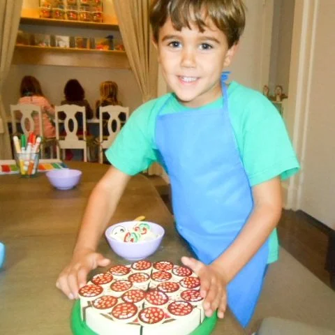 A young boy smiling while decorating a pizza-shaped cake with pepperoni slices in a home kitchen.