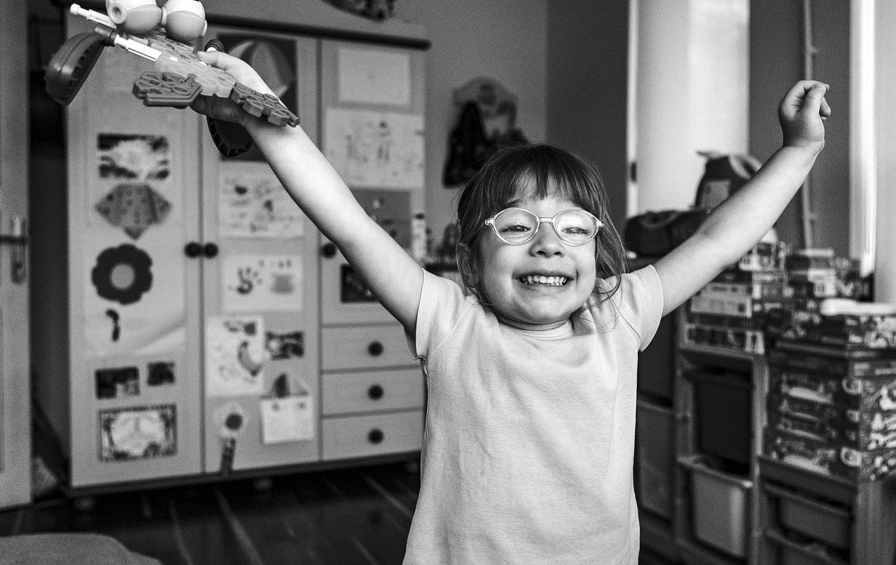 A smiling young girl with glasses raising her arms in a playroom filled with toys and furniture.