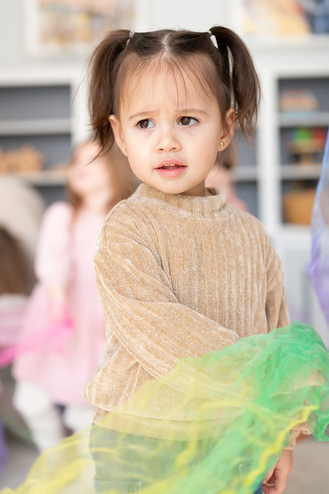 A young girl with brown hair in pigtails, looking confused or worried, wearing a tan long-sleeve shirt and standing in a room with other children.