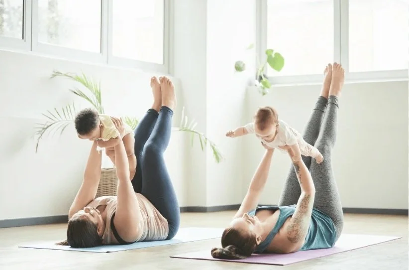 Two women doing yoga with children on a bright, airy room with large windows and plants
