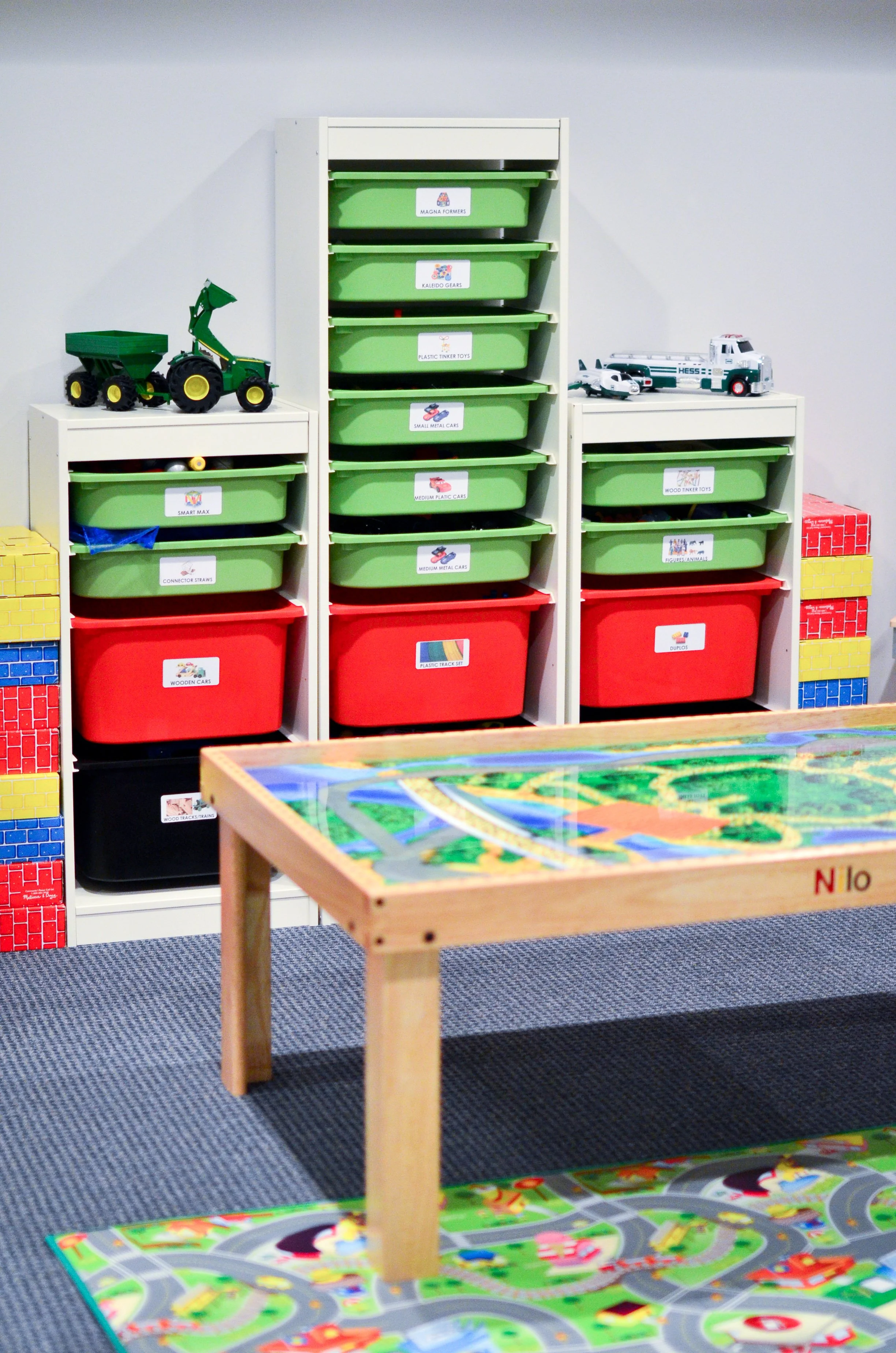 Toy storage shelves with labeled plastic bins containing toy vehicles and accessories, a play table with a city-themed play mat, and building blocks stacked on the sides of the shelves.