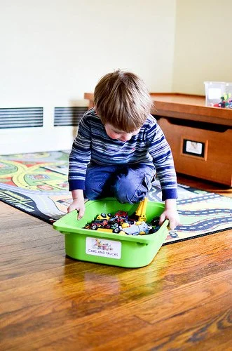 A young boy in a striped shirt playing with toy cars and trucks in a green plastic bin on a wooden floor.