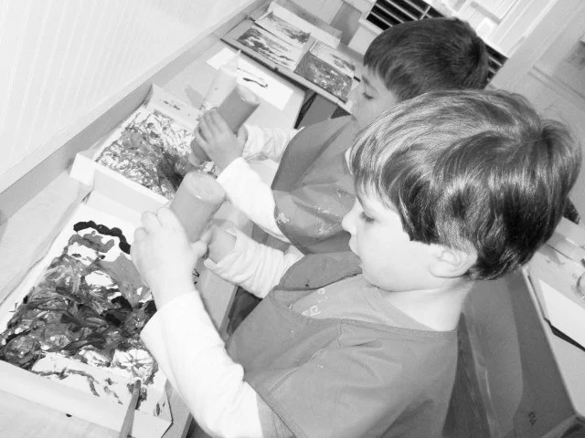 Two young boys at a craft table with bottles of paint and a tray of painted objects.