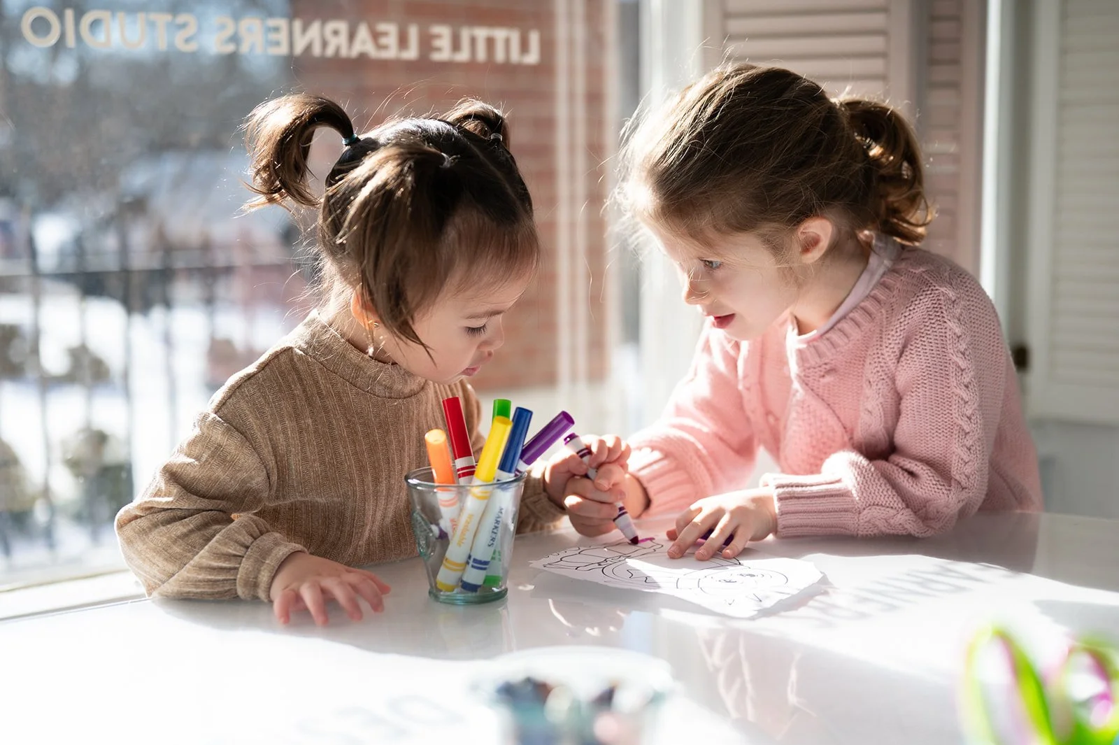 Two young girls sitting at a table, drawing with colored markers on a piece of paper. One girl is wearing a brown sweater, and the other is wearing a pink sweater. Bright sunlight is streaming in through a window behind them.