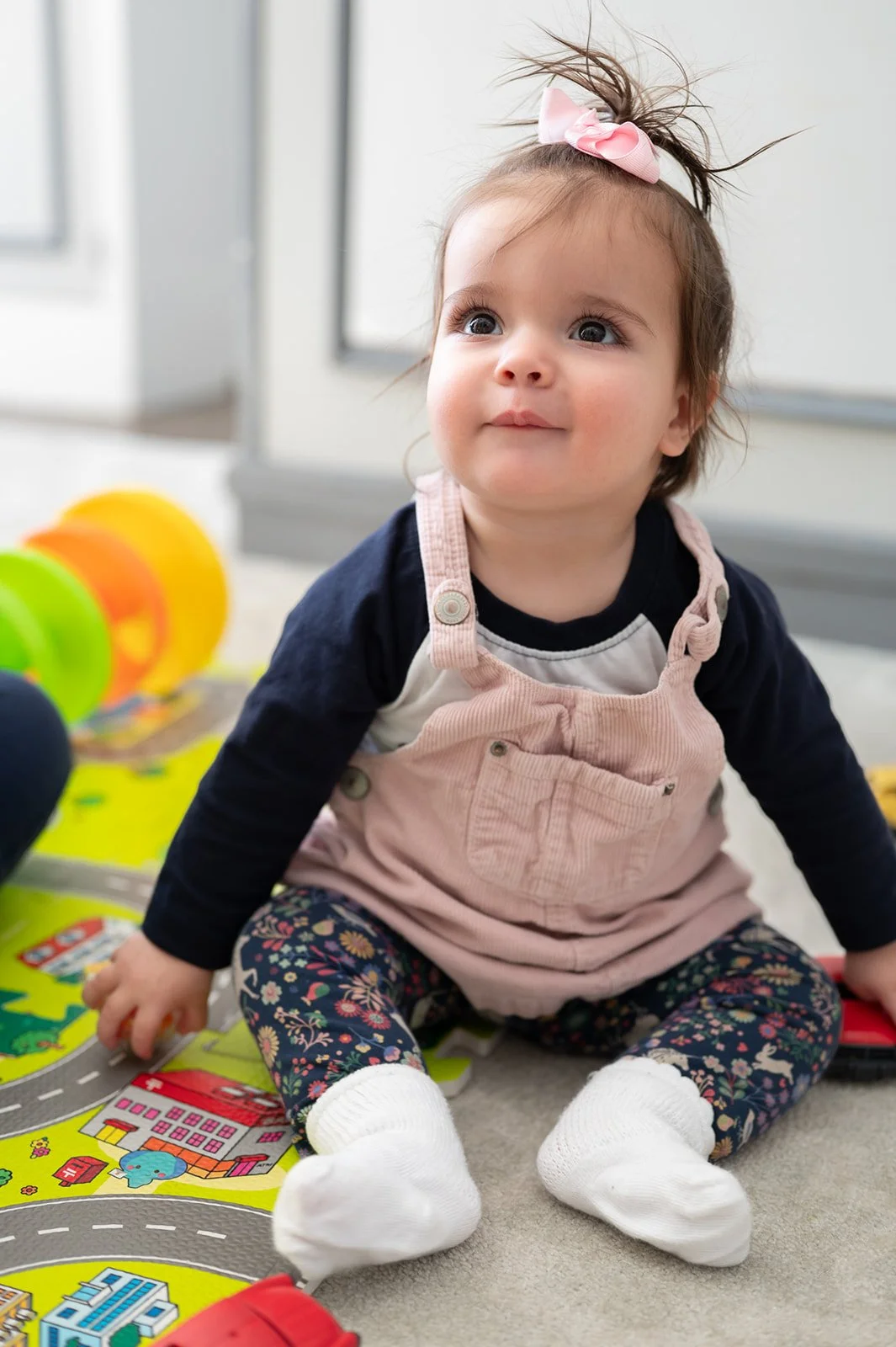 A young girl sitting on a colorful play mat, looking slightly upward with a curious expression, wearing a pink overall, floral leggings, white socks, and a pink bow in her hair.