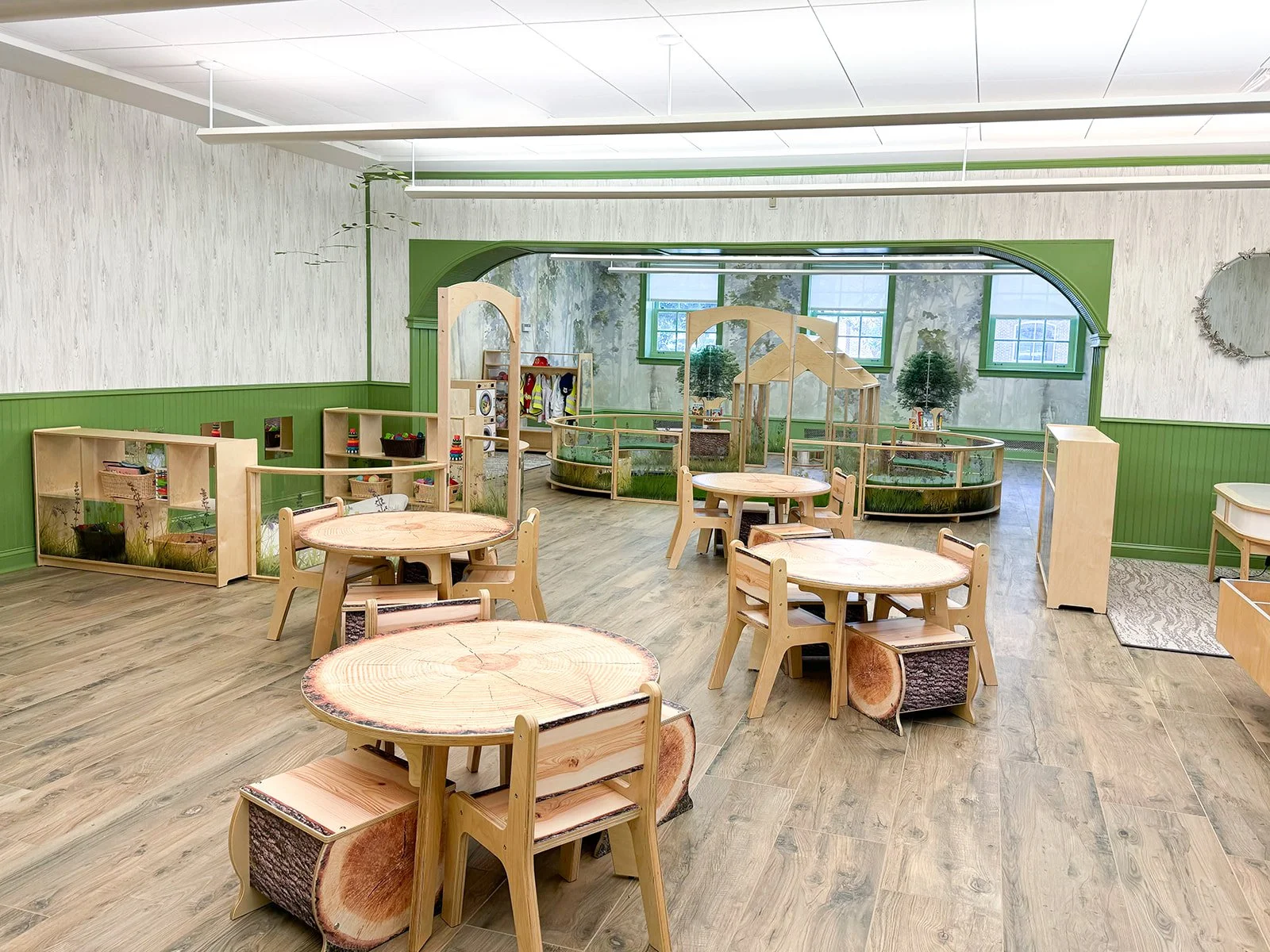 A spacious children's playroom featuring wooden tables and chairs made of tree slices, wooden partitions with nature-themed designs, and a partially enclosed play area with miniature trees and a glass railing, all decorated in green and natural wood tones.