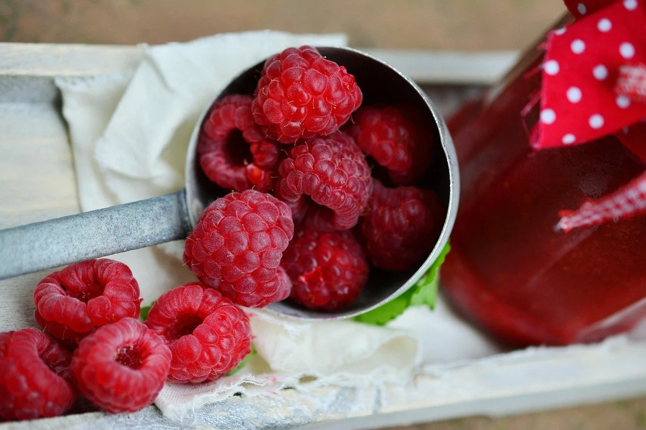 Fresh red raspberries in a metal scoop and on a white cloth with a jar of raspberry jam in the background.