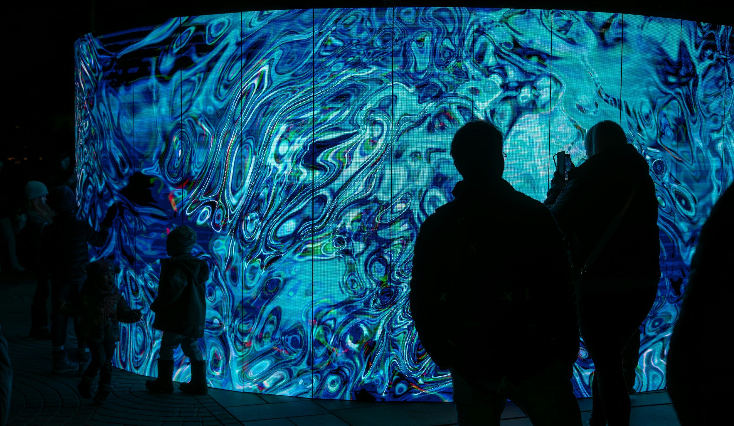 People gathering around a large, curved digital art display with swirling blue, green, and black patterns at night.