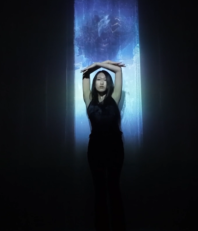 A woman standing with her arms crossed above her head in front of a digital or light projection on a wall, with a dark background.