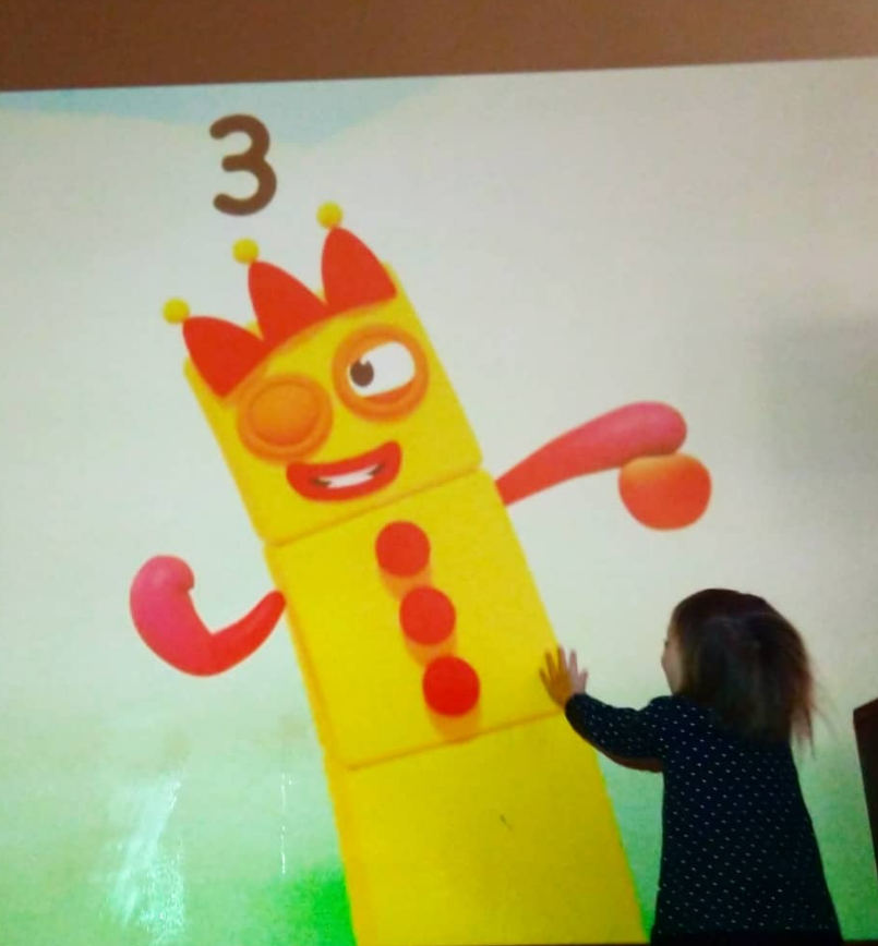 Child touching a colorful cartoon character on a wall, resembling a yellow dice with a face, arms, and a crown.