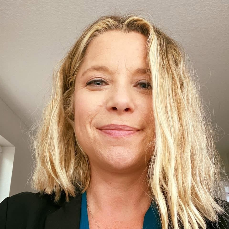 A woman with blonde, wavy hair smiling in an indoor setting.