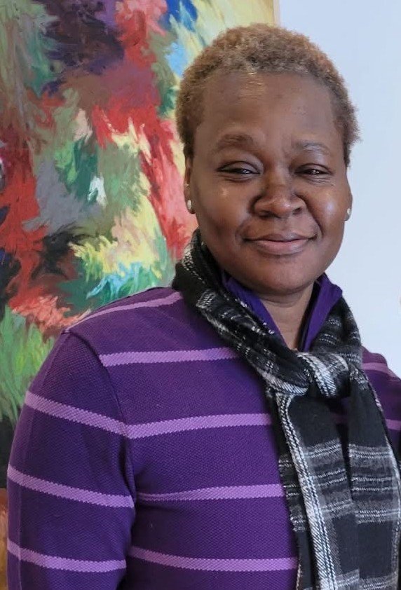 A middle-aged Black woman with short curly hair, wearing a purple striped sweater and a black and white checkered scarf, standing in front of a colorful abstract painting, smiling gently.