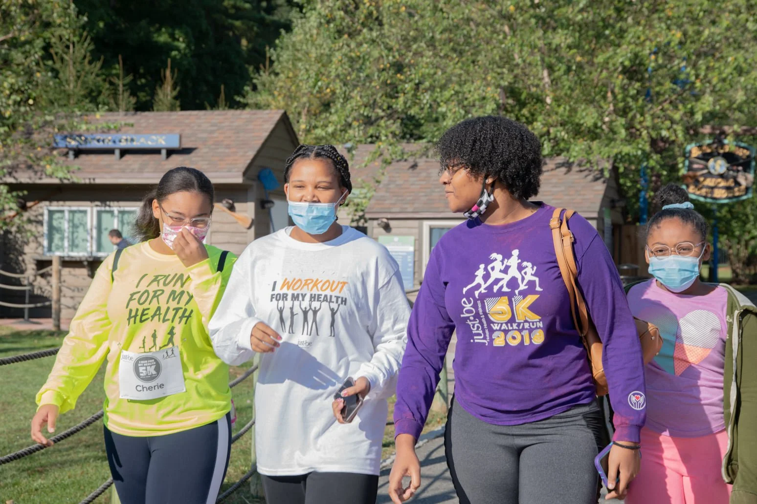 Four women, all wearing face masks, walking outdoors near a wooden building and trees, during a community walk event.
