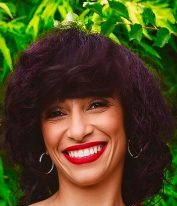 Close-up of a woman with curly dark hair smiling, with green leaves in the background.