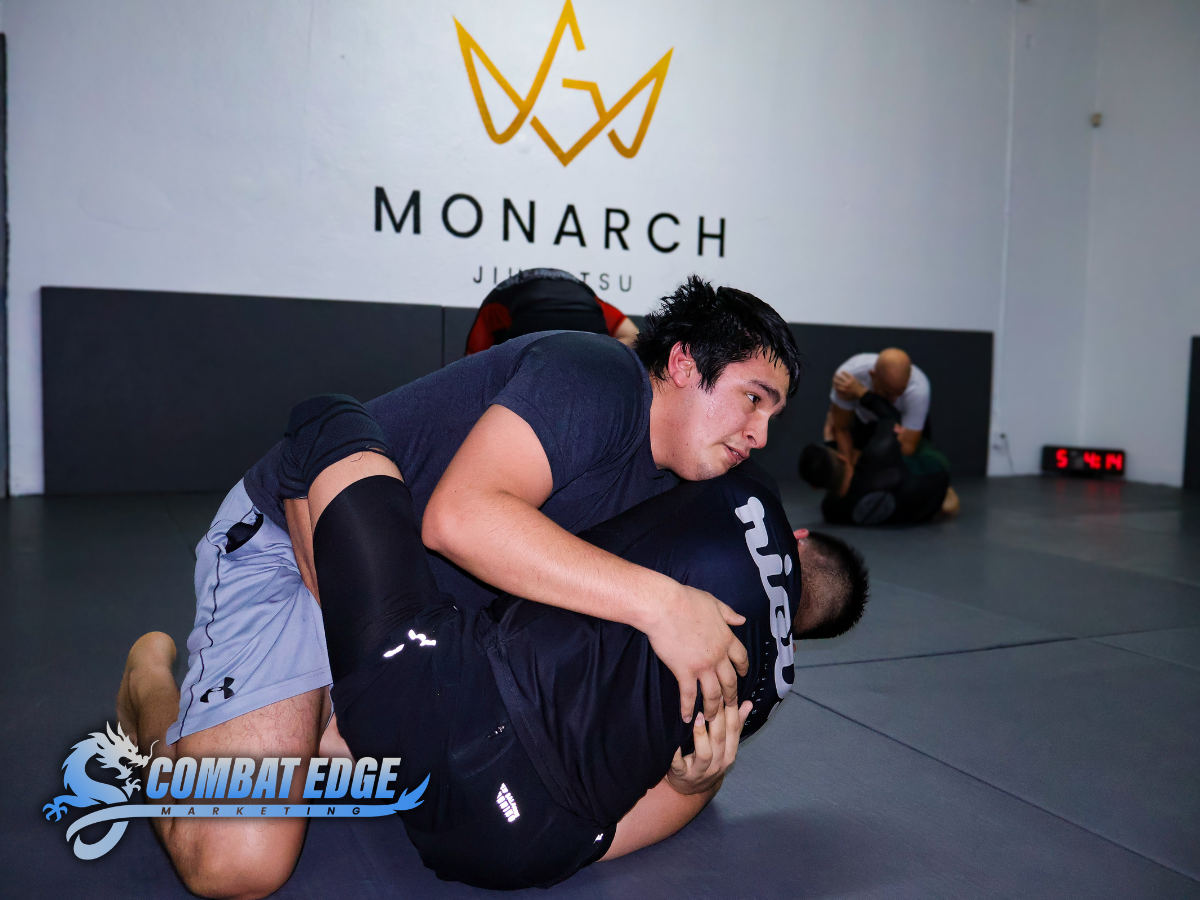 Two men practicing Brazilian Jiu-Jitsu on a mat at Monarch Jiu Jitsu gym. One man is on top, controlling his opponent with a grip around the neck and arm, while the other is on his back attempting a guard position. There are other practitioners train
