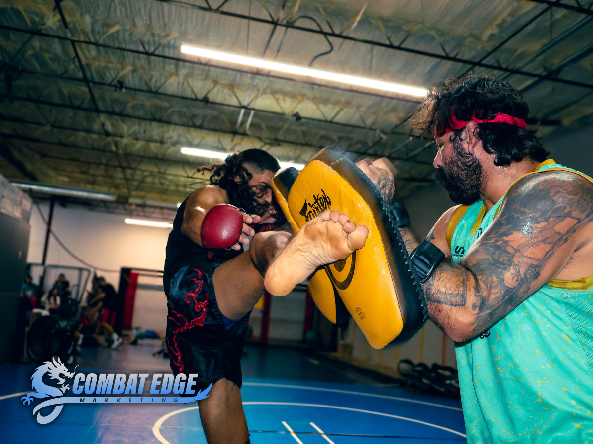 Two men practicing martial arts in a gym: one is sparring with a focus mitt held by the other, who is wearing red gloves and has tattoos, a headband, and a sleeveless shirt.