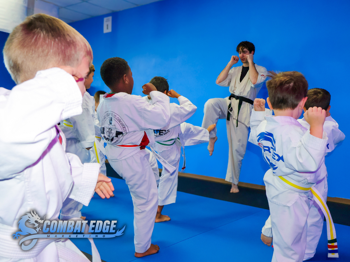 Martial arts instructor demonstrating kicks to children in martial arts uniforms in a training room.