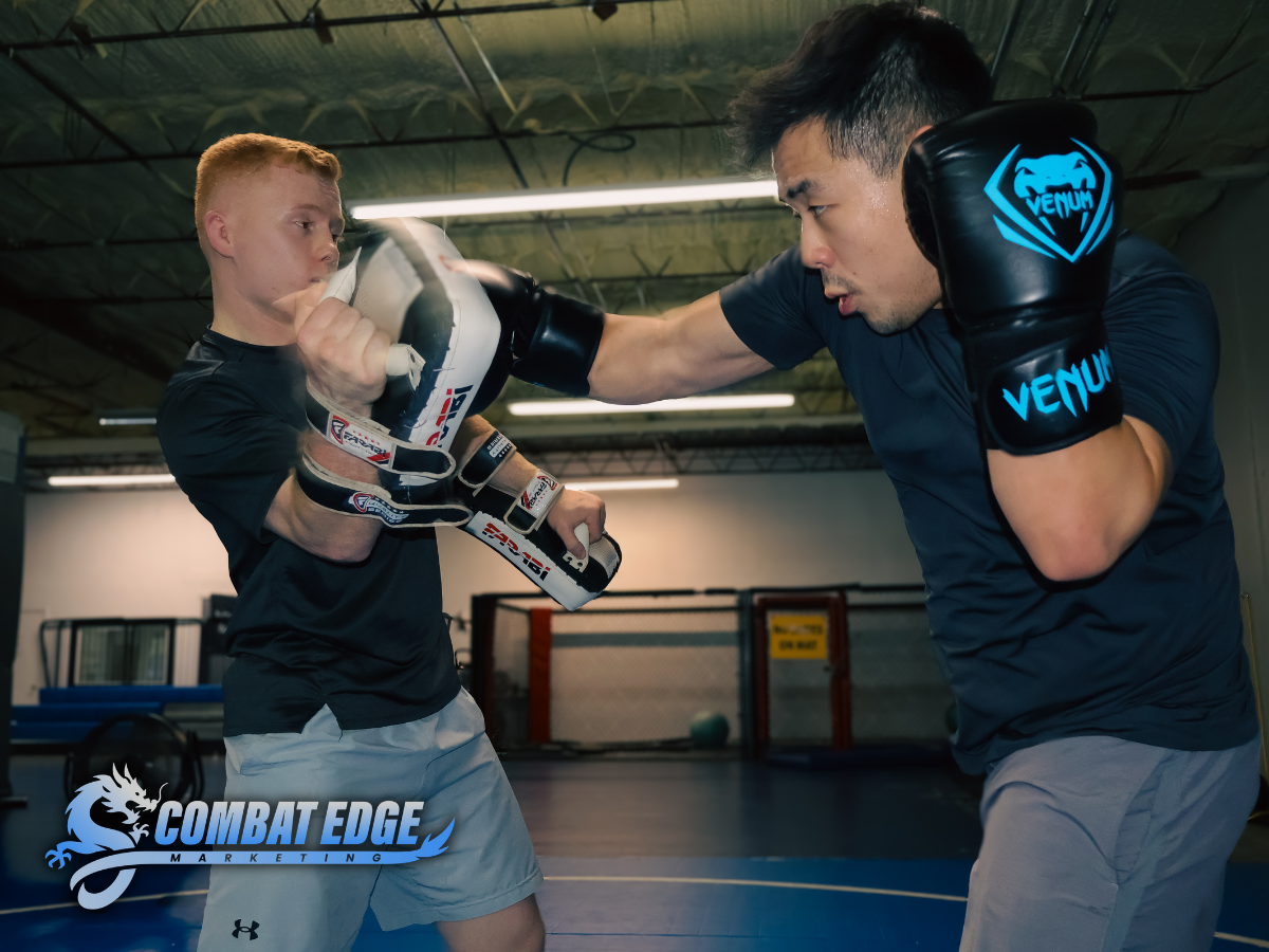 A young male in black shirt and shorts practicing boxing with an instructor in gloves inside a gym.