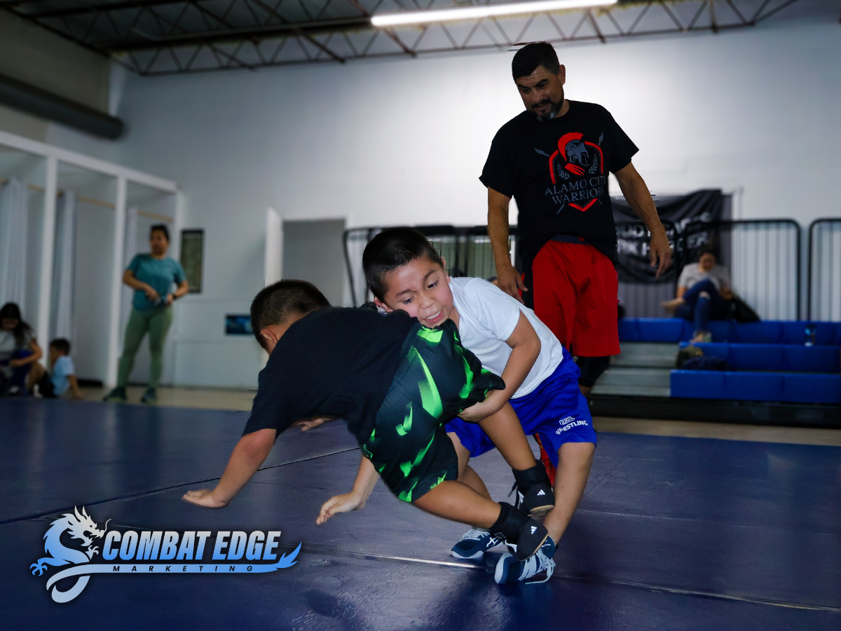 Two young boys wrestling on a mat in a gym with an adult supervising. Background shows other children and spectators, with a Compassion Edge Marketing logo in the bottom left corner.