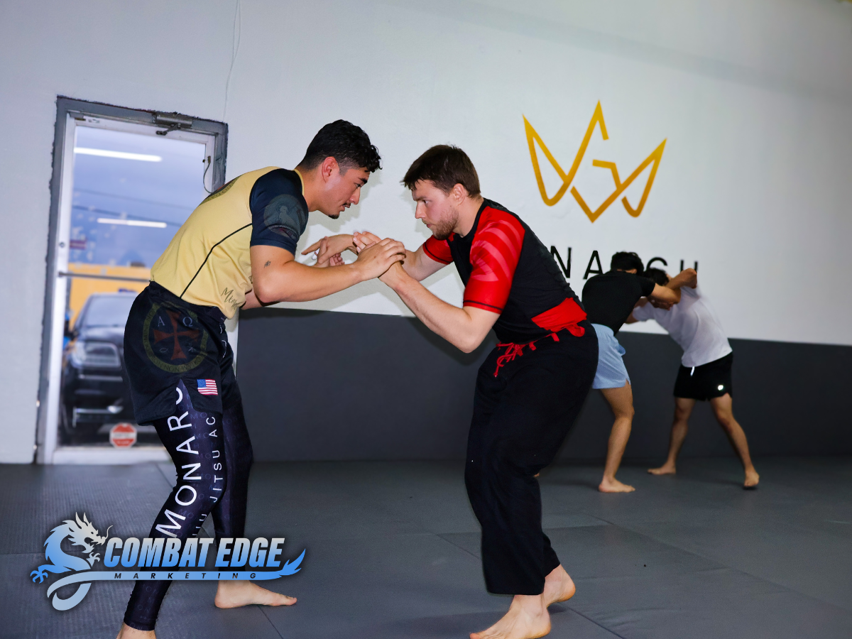 Two pairs of people practicing martial arts in a gym, with the two in the foreground engaged in a grappling drill, and two in the background practicing self-defense techniques. The gym has a logo on the wall and the words "COMBAT EDGE" and "MONARCH" 