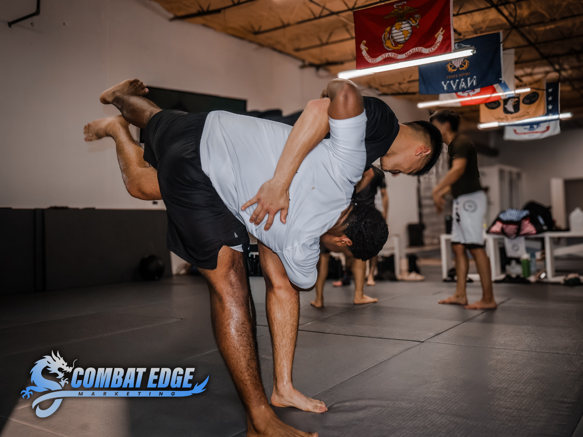 Two men practicing Brazilian Jiu-Jitsu in a martial arts gym, one in white and the other in black rash guard, grappling on padded mats with American flags and martial flags hanging from the ceiling.