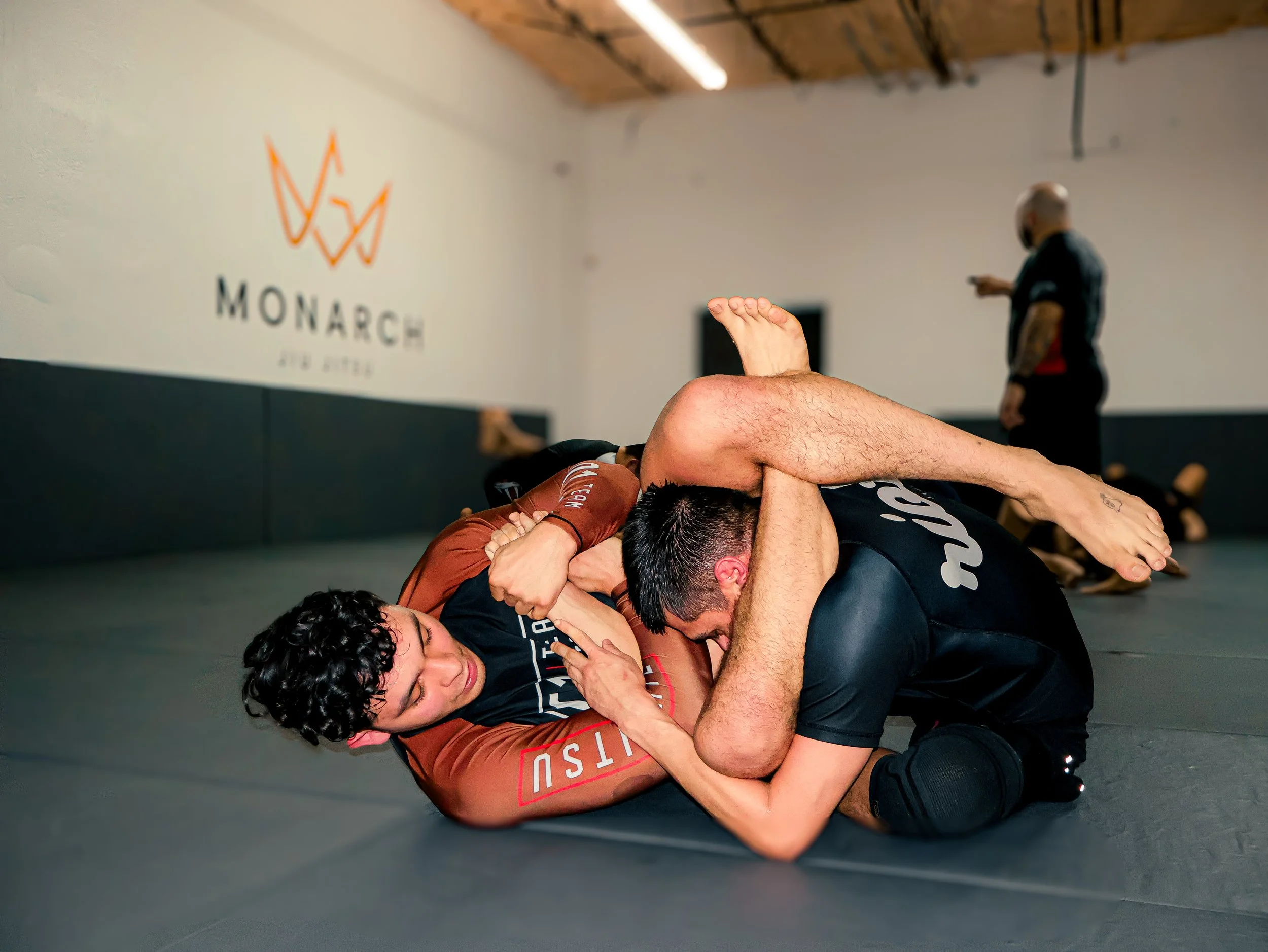 Two male Brazilian Jiu-Jitsu practitioners engaged in a grappling match on the mat inside a gym, with a coach or referee in the background observing.