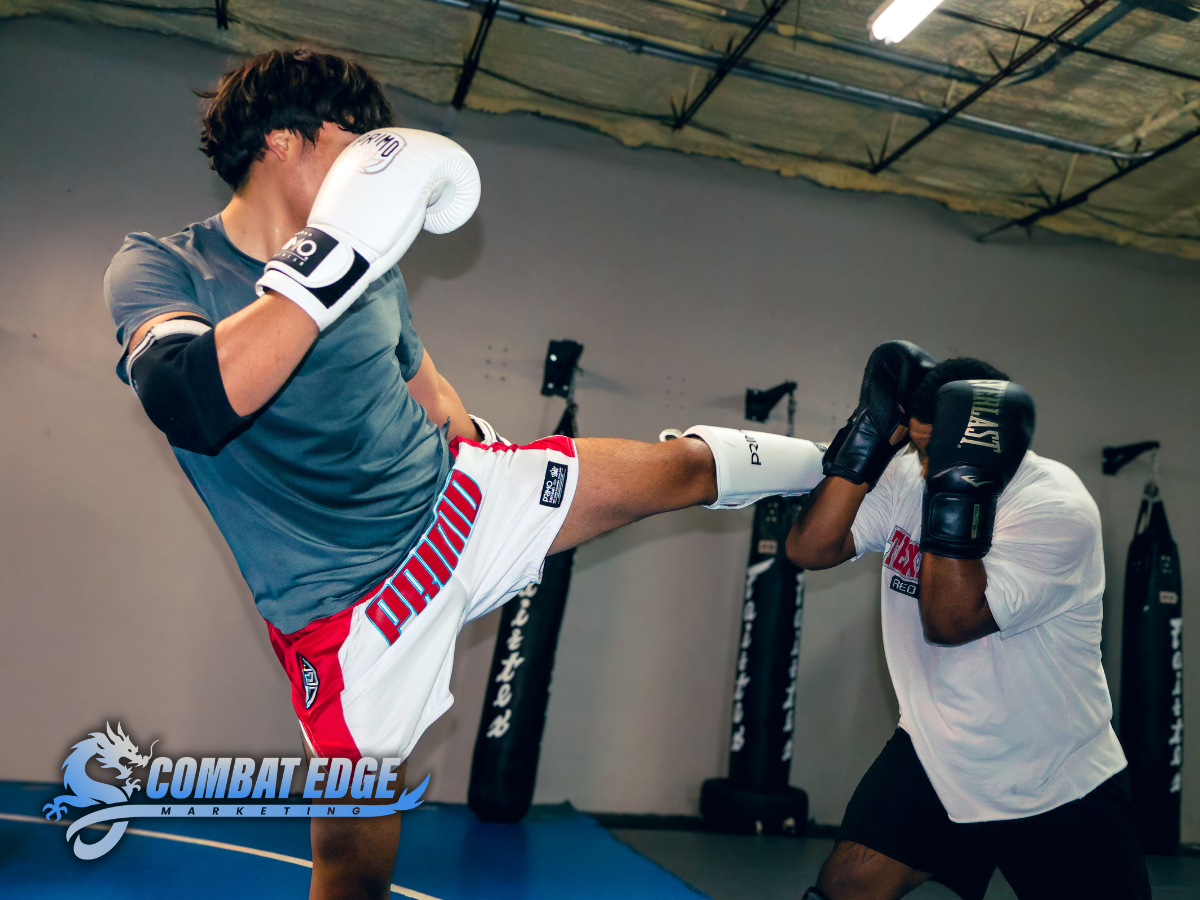 An MMA fighter performs a high kick targeting a training mitt held by a trainer in a gym, with punching bags in the background.