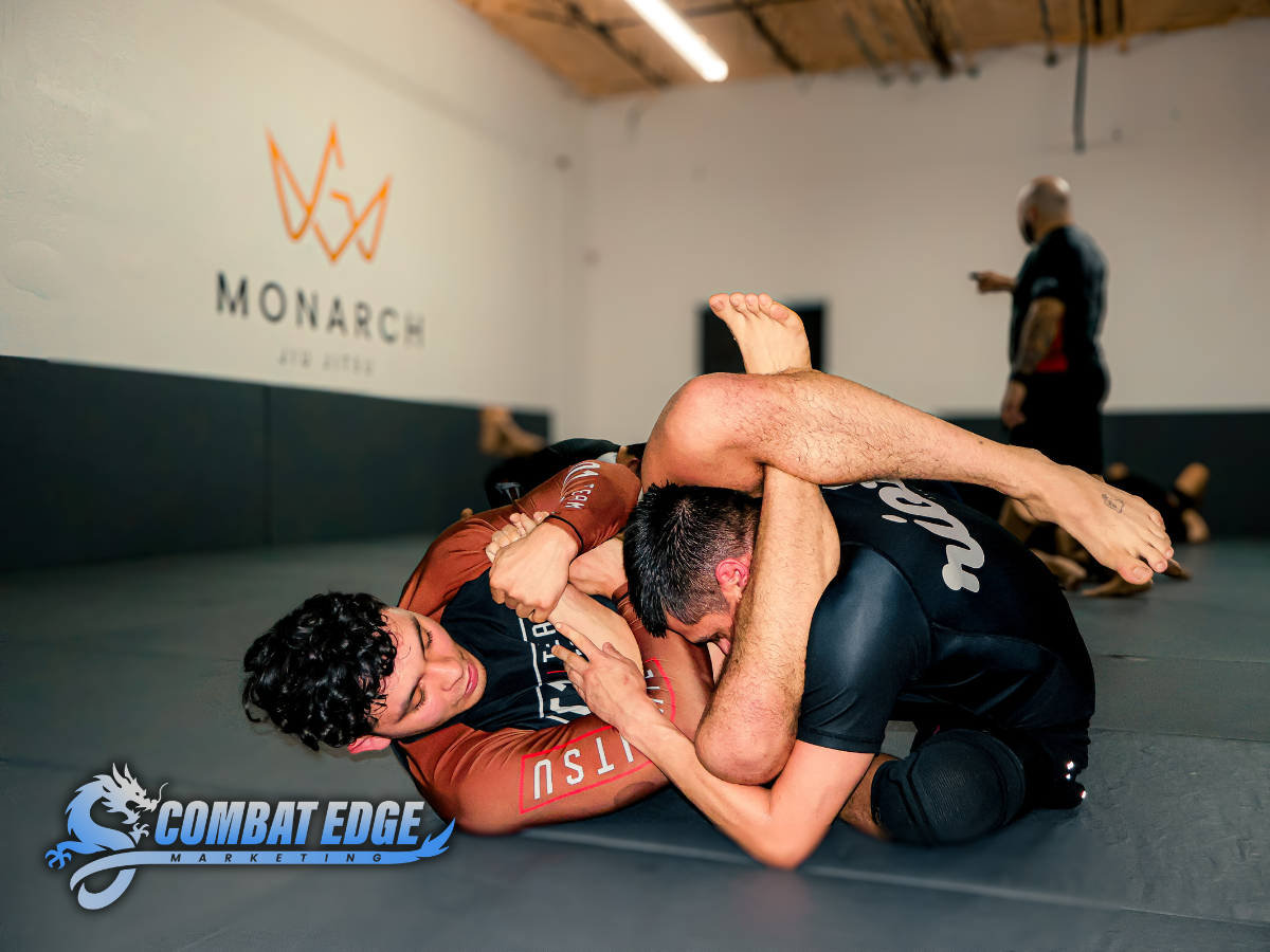 Two men practicing Brazilian Jiu-Jitsu on a mat in a gym, with one man in a black shirt and the other in a brown rash guard, fighting in a grappling position. In the background, a referee or instructor is observing, and there are other practitioners 