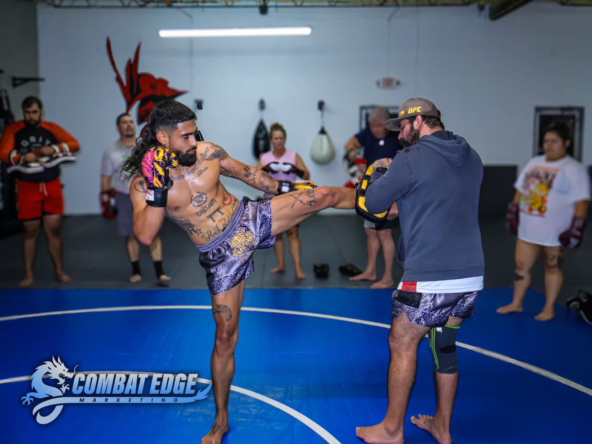 A mixed martial arts fighter practicing a kick during training with an instructor in a gym, with several people observing in the background.