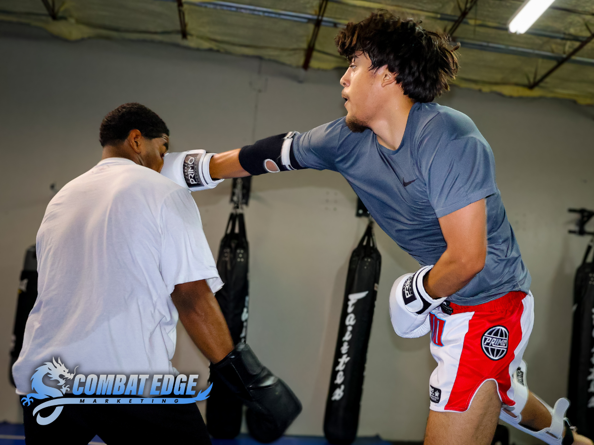 Two men training in a gym, one wearing boxing gloves and practicing punches, the other wearing boxing mitts holding pads for training, with boxing bags hanging in the background.