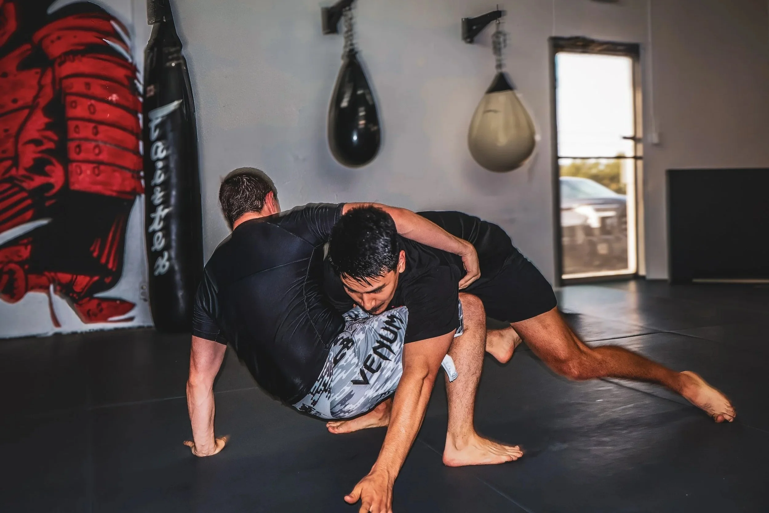 Two men practicing Brazilian Jiu-Jitsu on a mat in a gym, with punching bags hanging on the wall and a door with a window in the background.