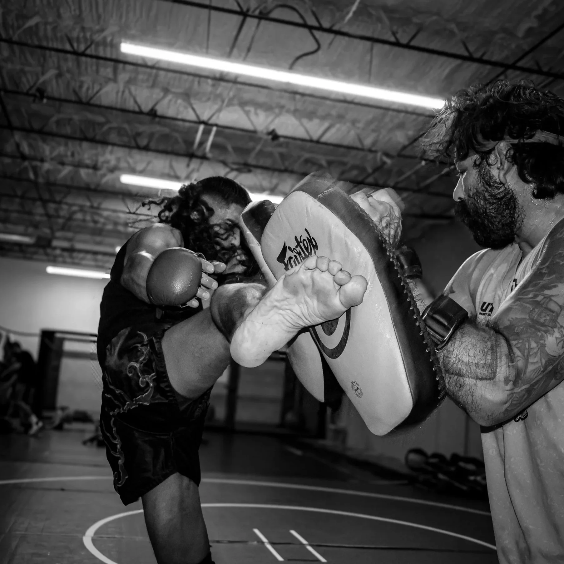 Two men practicing martial arts in a gym, focused on a padded kicking shield. One man is delivering a kick to the shield while the other holds it. The setting appears to be a training area with overhead lighting and a wooden ceiling.
