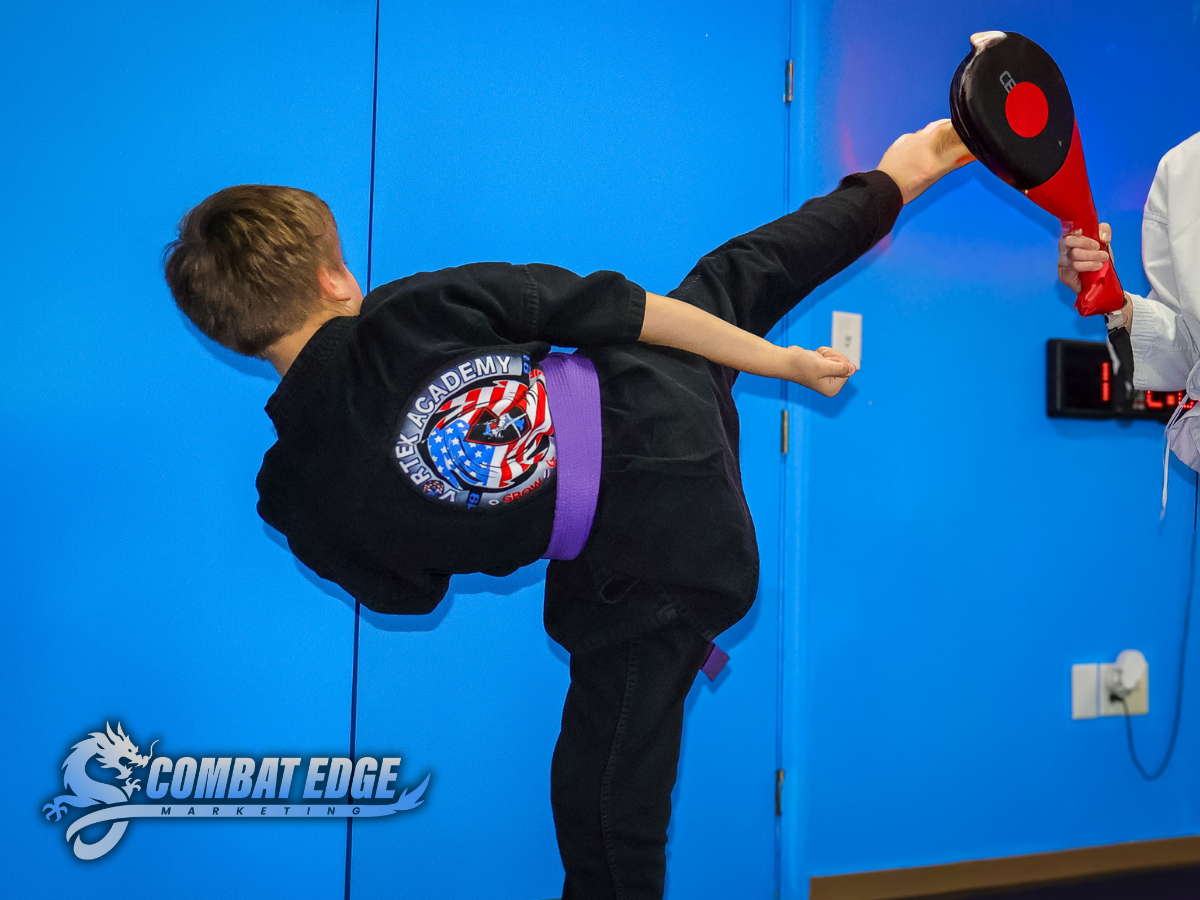Person practicing martial arts high kick against a blue wall, wearing a black gi with a purple belt and a patch on the back, aided by a trainer holding a kicking pad.