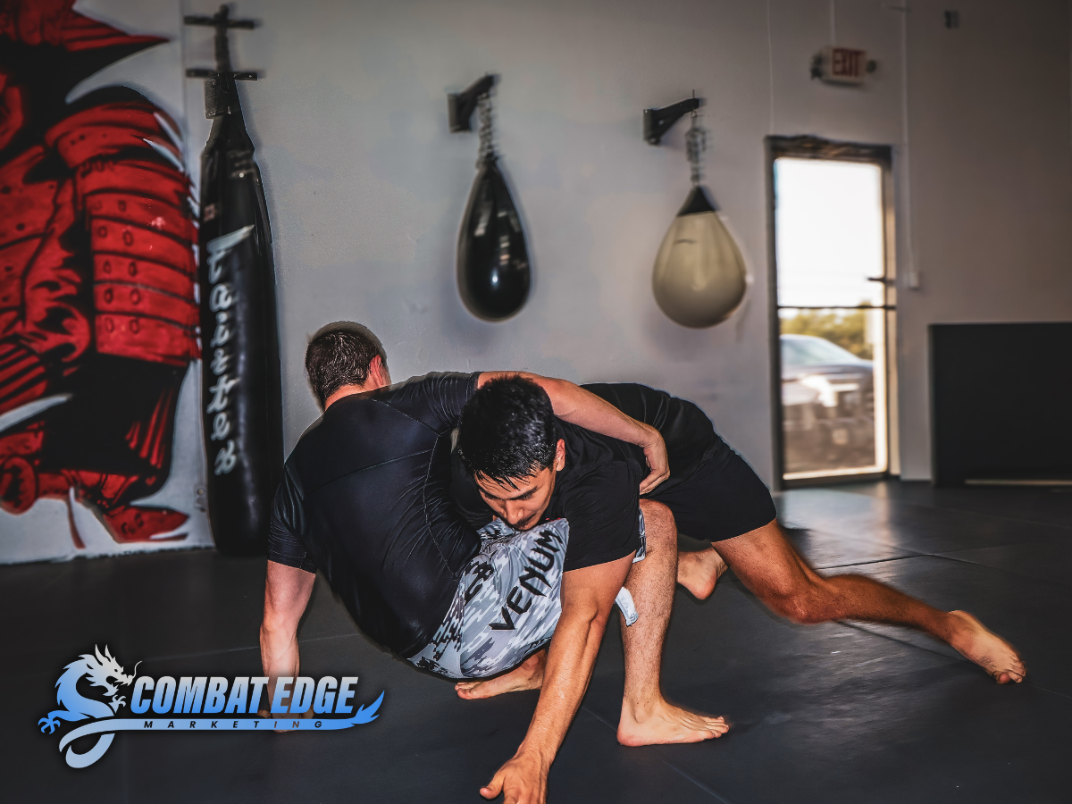 Two men practicing Brazilian Jiu-Jitsu on a black mat in a gym. One man is on top, gripping the other's arm, while the other man is on all fours. Gym equipment and a punching bag are visible in the background, with the gym's logo "Combat Edge" in the