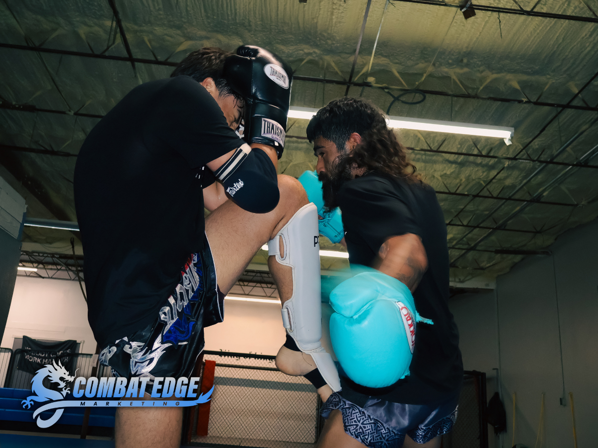 Two men sparring in a martial arts gym. One is wearing black gloves, shin guards, and shorts, while the other is wearing blue boxing gloves and shorts. The man with the gloves is delivering a knee strike to the other's midsection.