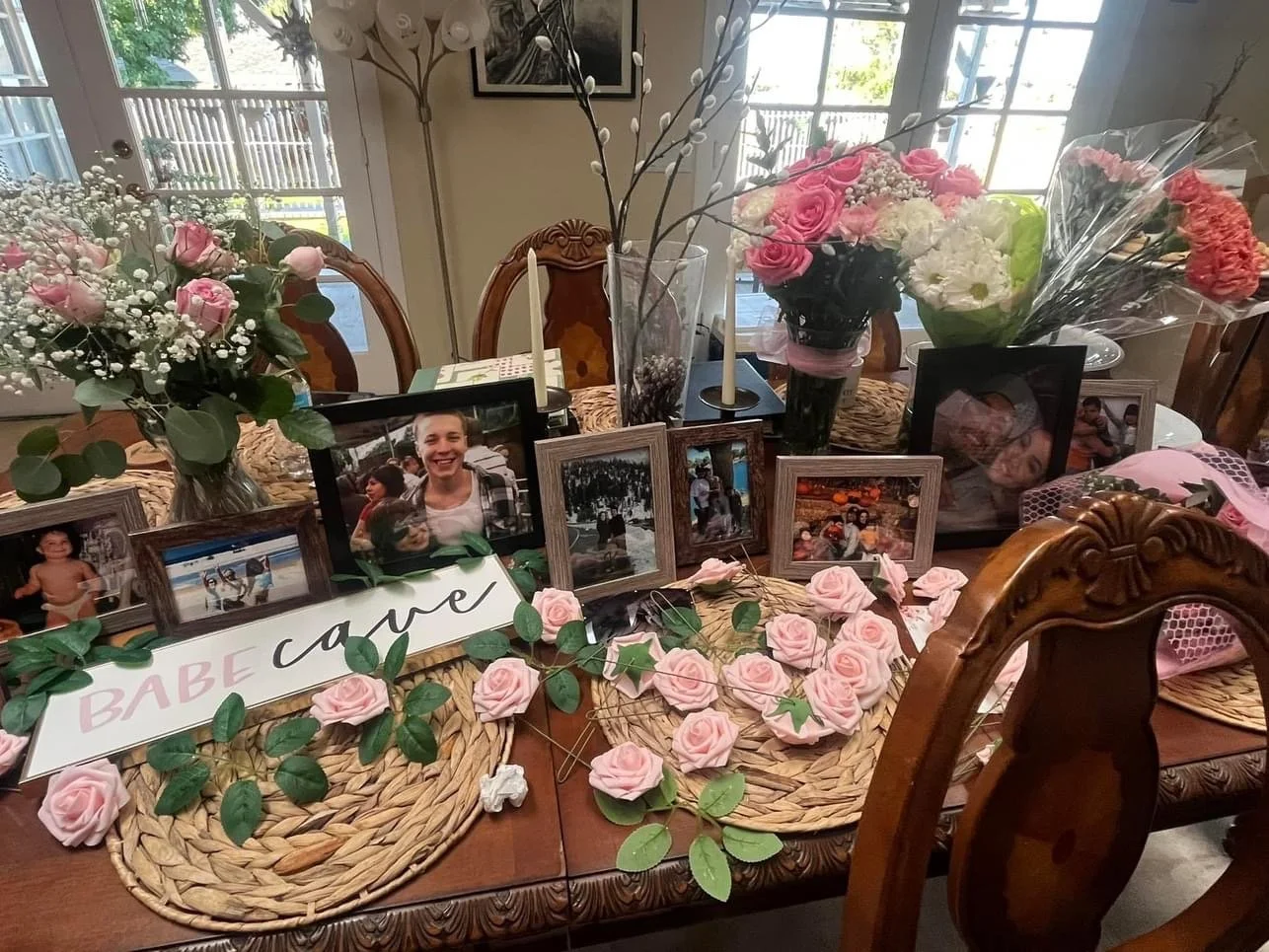 A dining table decorated with pink and white floral arrangements, framed photographs, a sign that reads "BABE cave," and decorative pink roses and greenery.