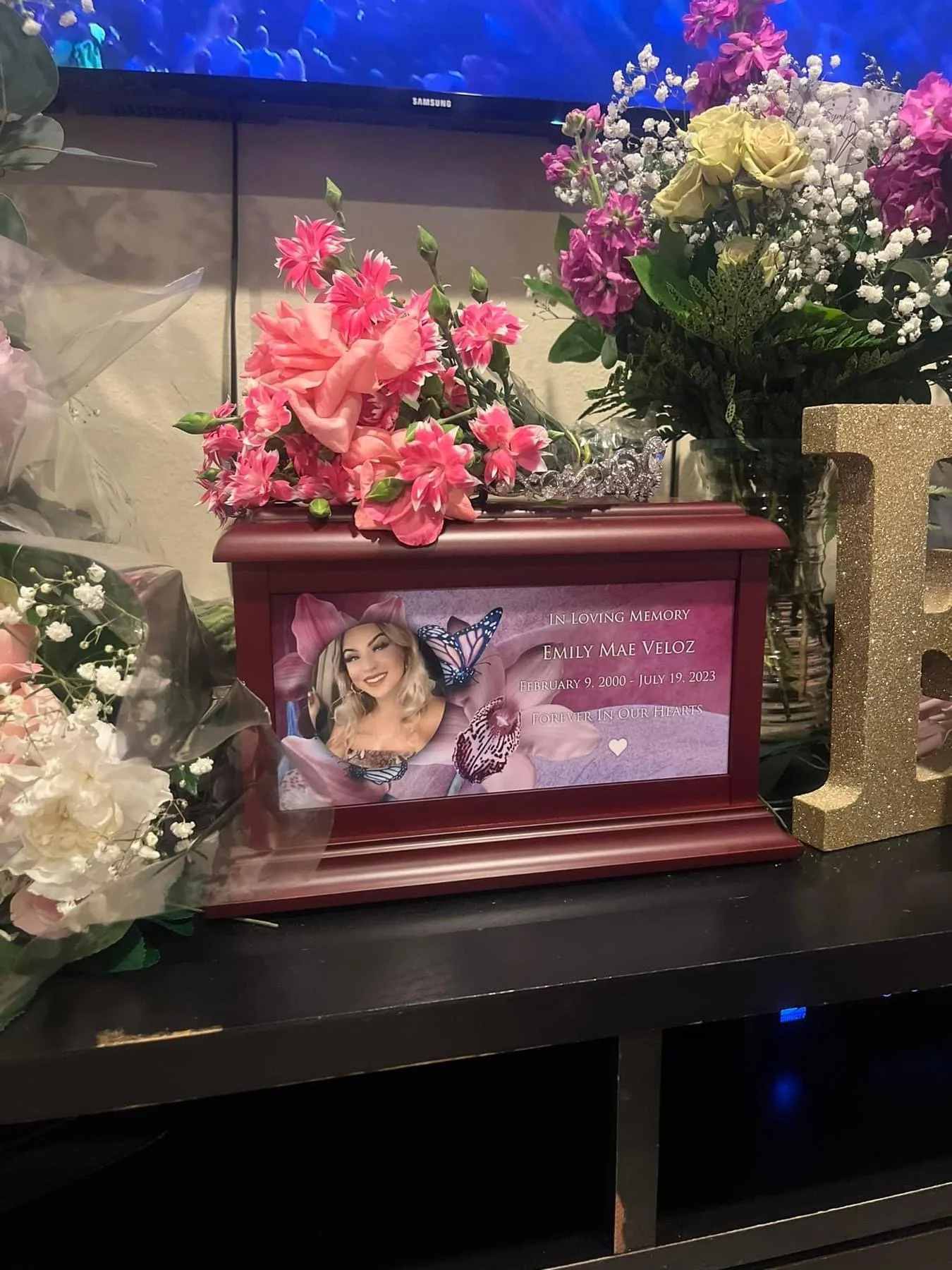 Memorial display with pink and white flowers, a framed photo of a smiling young woman with blonde hair and butterfly decorations, and a gold glitter letter 'L' on a black surface.