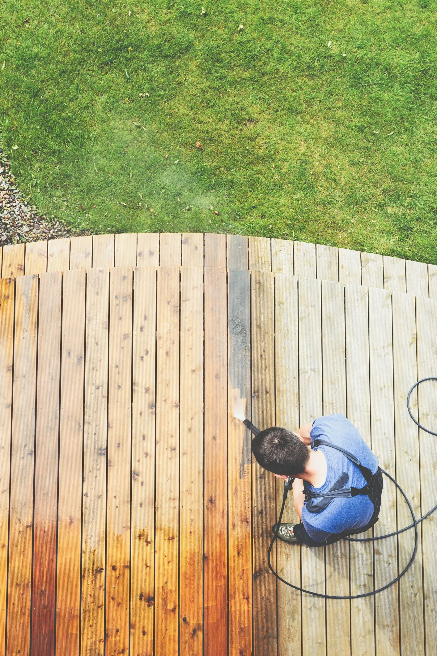 A person pressure washing a wooden deck near grass on a sunny day.