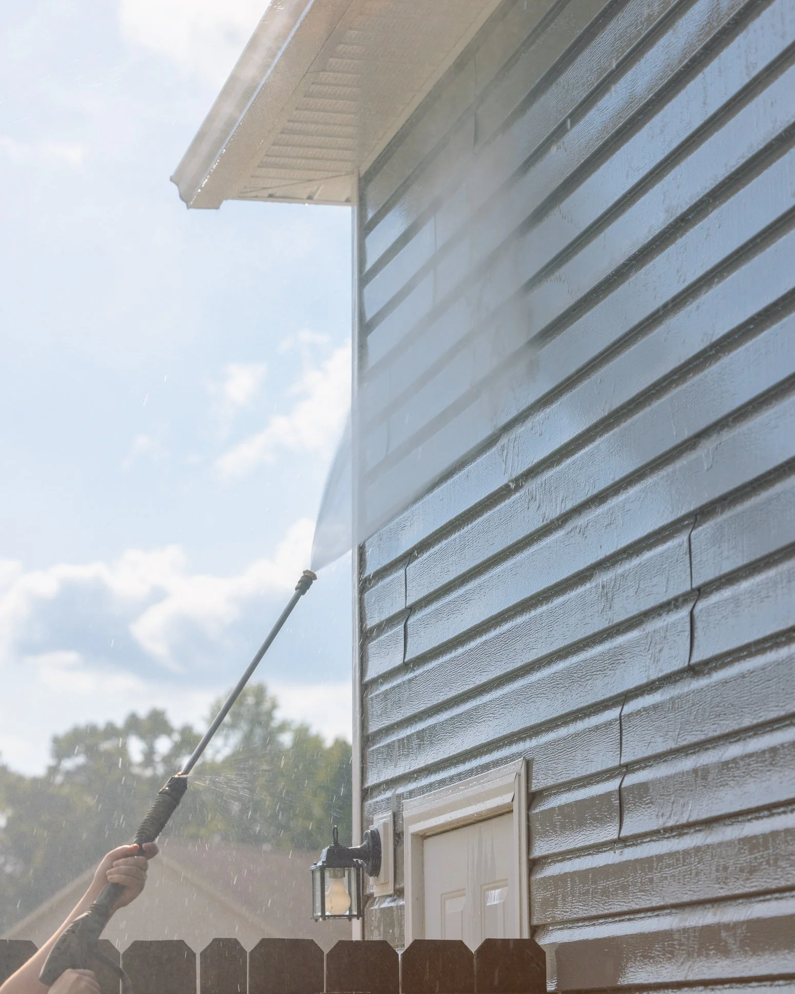 Person spray washing the side of a house with a pressure washer sunlight in the background