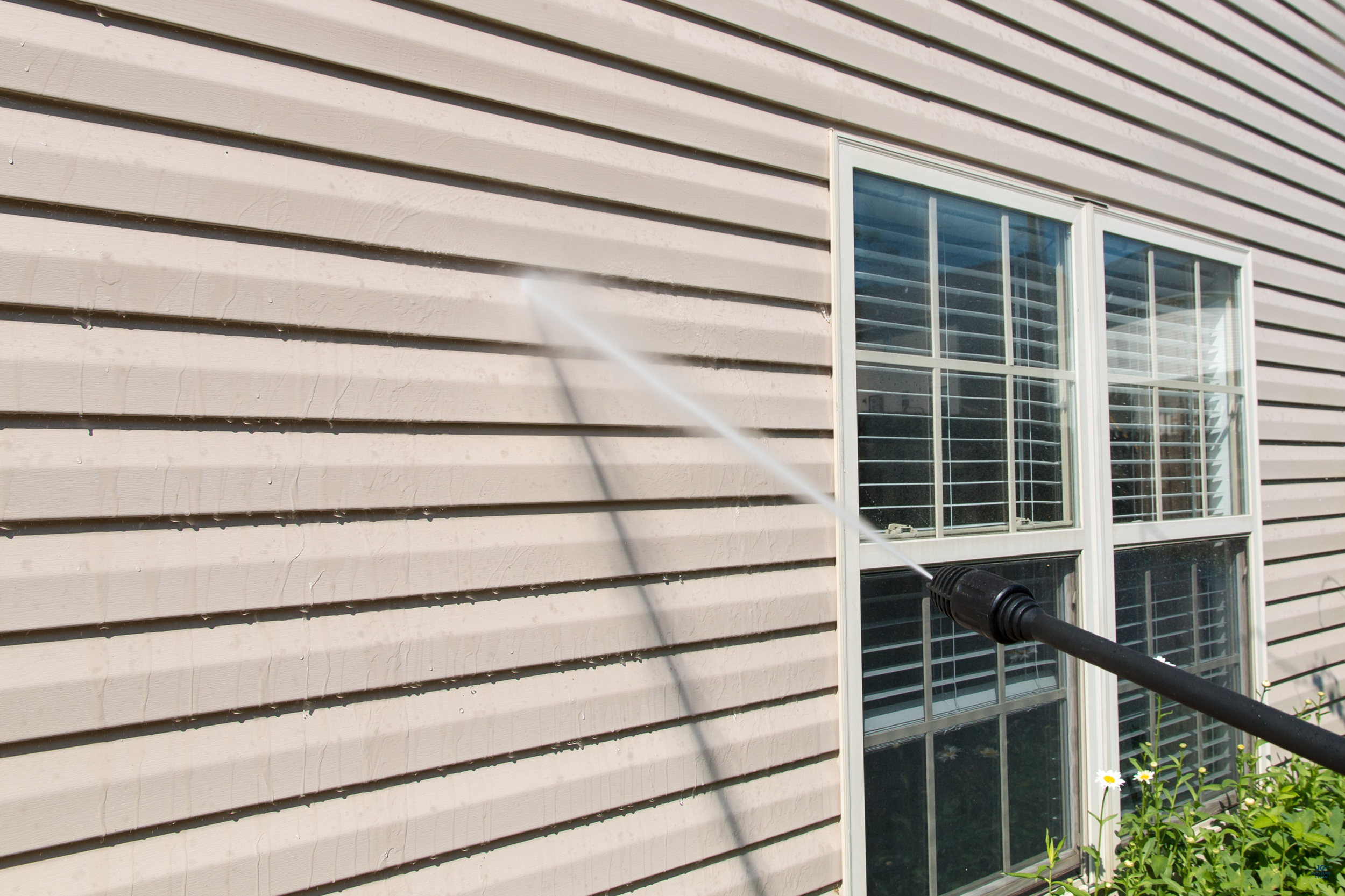 A garden hose spray nozzle is spraying water on the exterior wall of a house with beige vinyl siding and a window.