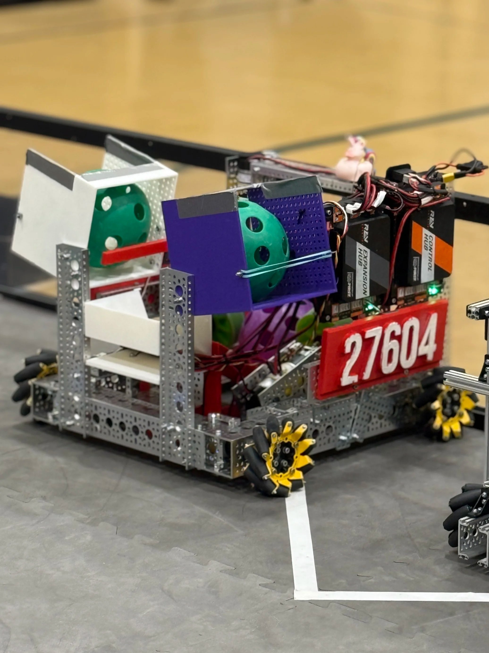 A robot made of metal parts with wheels, sensors, and electronic components, participating in a robotics competition on a gymnasium floor.