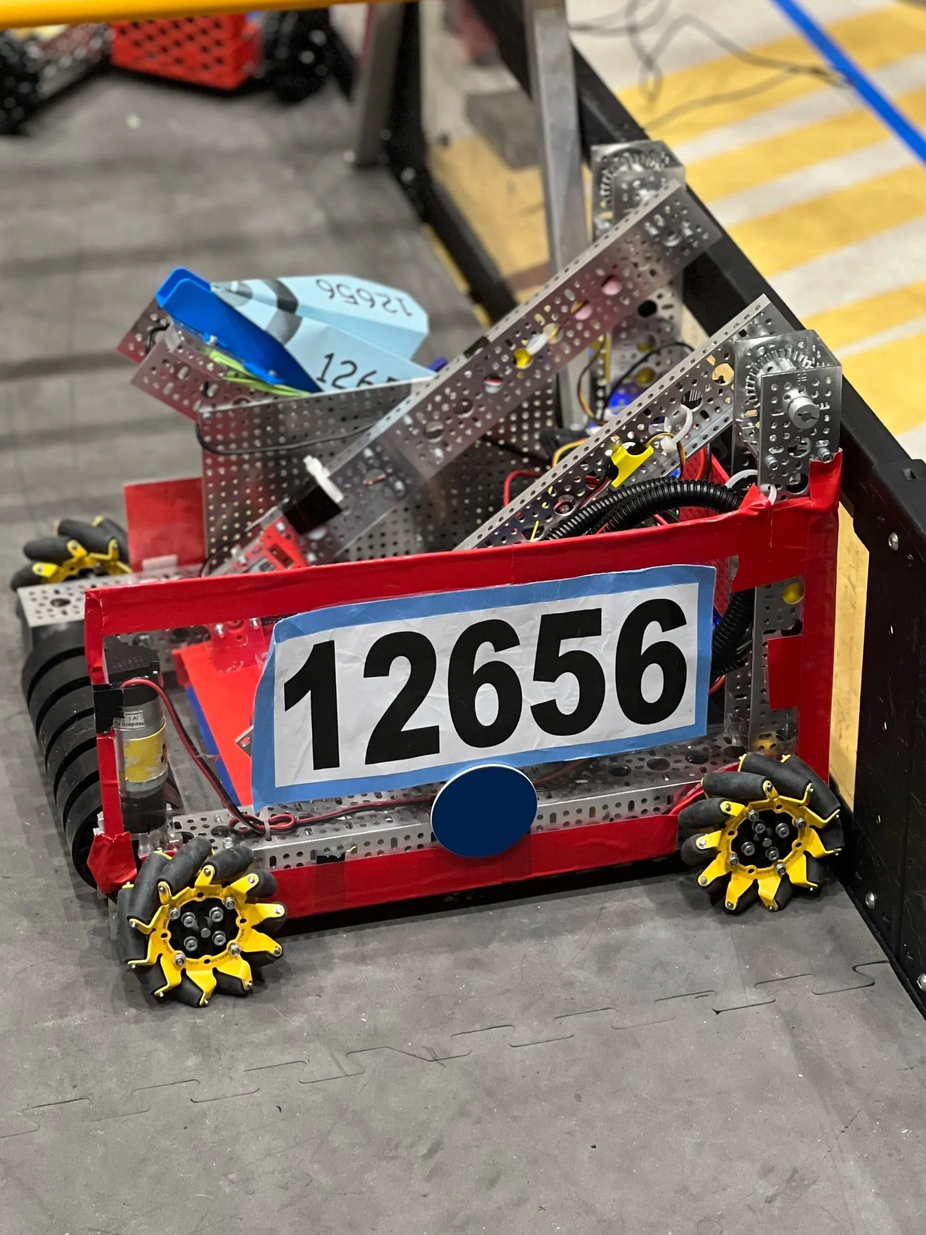 A robot constructed from metal parts, with a red frame, yellow roller wheels, and a blue circular element. It has a large white label with the number 12656 on the front. The robot appears to be part of a robotics competition or project.