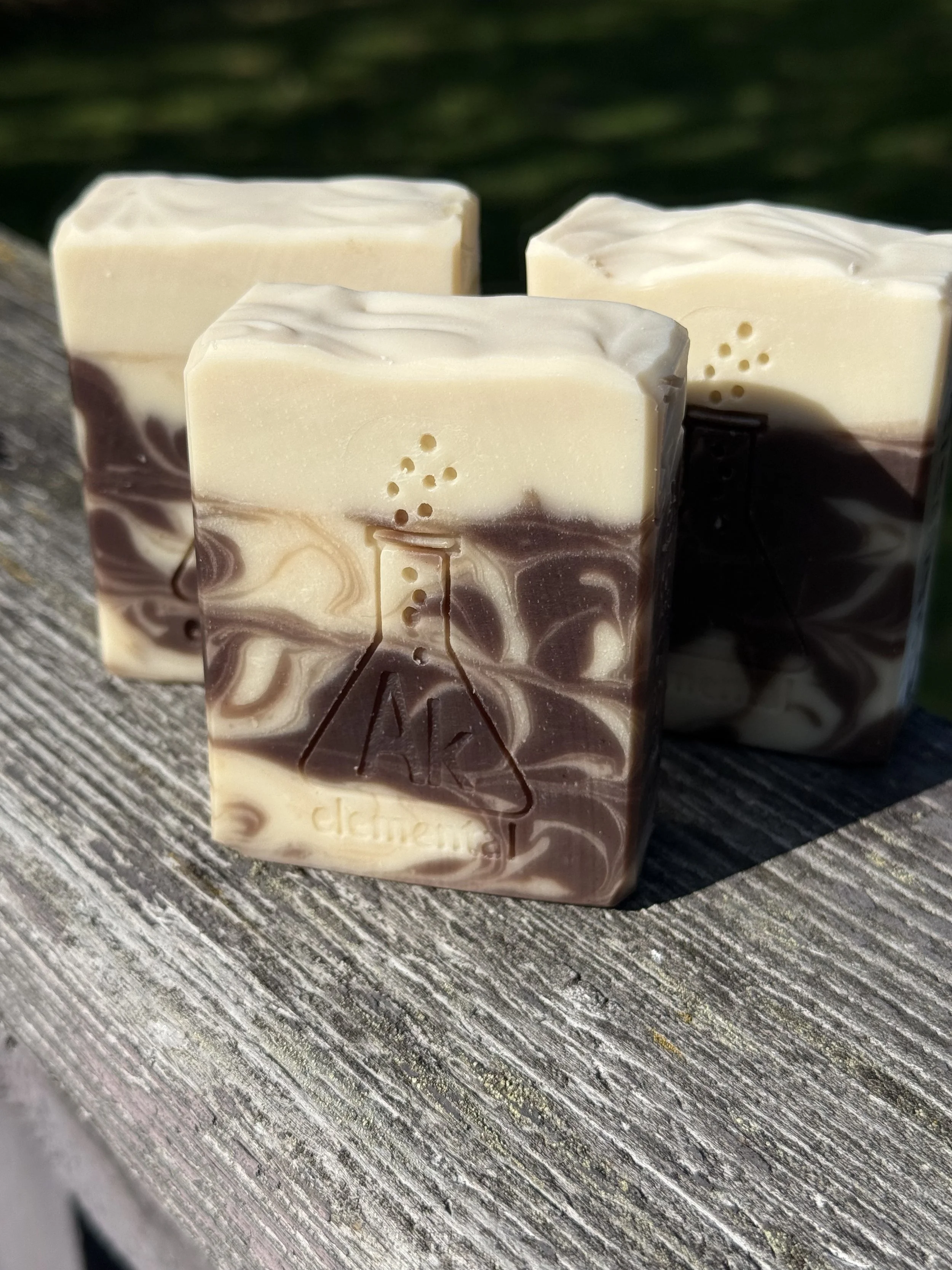 Three bars of soap with brown and cream swirls and cream top sitting on a weathered board. AK Elemental logo of an erlenmeyer flask is stamped on the soaps.